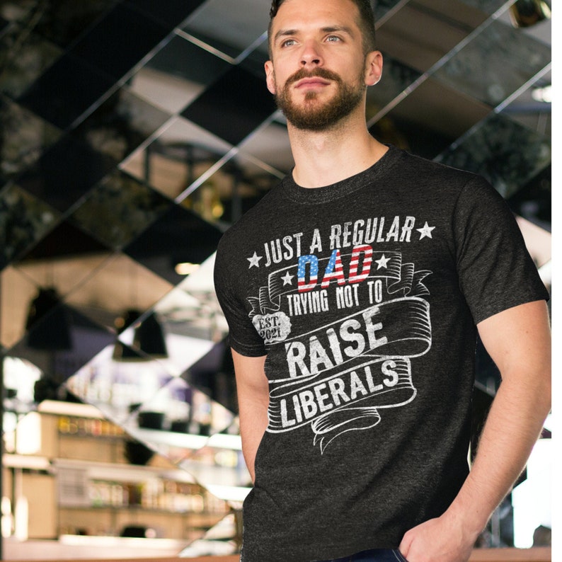 Op de afbeelding: Donkergrijs t-shirt met de tekst "JUST A REGULAR DAD trying not to RAISE LIBERALS." Het woord "DAD" heeft een rood-blauw Amerikaans vlagontwerp. Het shirt heeft ook de tekst "EST. 2021."