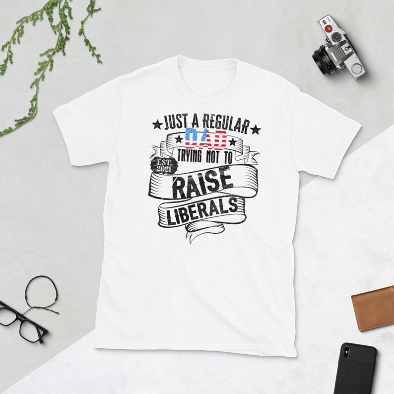Op de afbeelding: Wit T-shirt met de tekst "JUST A REGULAR DAD TRYING NOT TO RAISE LIBERALS" in zwarte en rode letters. Het woord "DAD" is ontworpen met een rood-wit-blauw vlagpatroon. Het shirt bevat ook "EST. 2021".