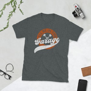 K&ouml;nnte beinhalten: Dunkelgraues T-Shirt mit dem Text "I'll Be In The Garage" in wei&szlig;er Schrift im Retro-Stil, mit einem orangefarbenen Kreis und einer Kolben-Grafik. Das Shirt besteht aus einem weichen Material.