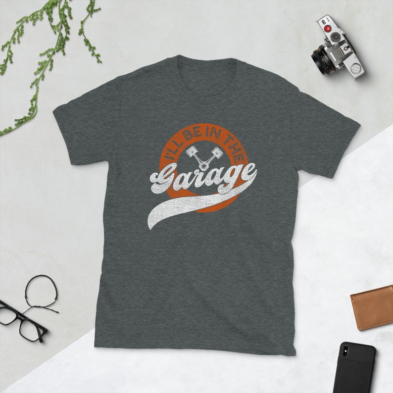 K&ouml;nnte beinhalten: Dunkelgraues T-Shirt mit dem Text "I'll Be In The Garage" in wei&szlig;er Schrift im Retro-Stil, mit einem orangefarbenen Kreis und einer Kolben-Grafik. Das Shirt besteht aus einem weichen Material.