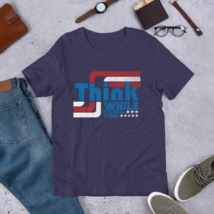 May include: A navy blue t-shirt with a red, white, and blue American flag design. The shirt has the text "Think While It's Still Legal" printed on it.