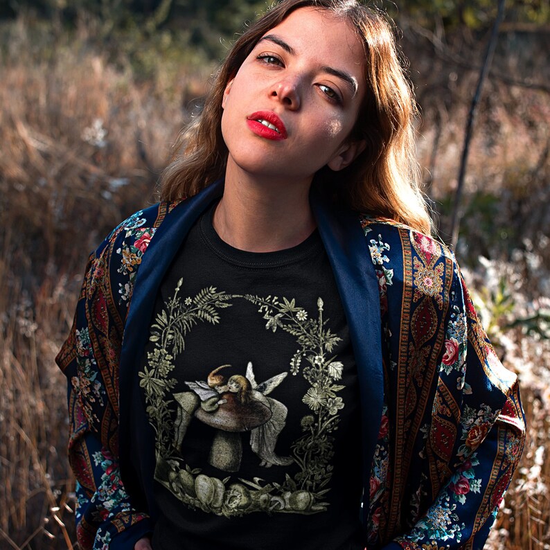 K&ouml;nnte beinhalten: Schwarzes Rundhals-T-Shirt mit einer detaillierten Illustration eines Pilz- und Feen-Designs in einem floralen Kranz. Das Model tr&auml;gt eine blau gebl&uuml;mte Jacke mit dunkelblauem Besatz. Der Hintergrund ist eine verschwommene Szene im Freien.