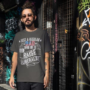 Op de afbeelding: Donkergrijs t-shirt met de witte tekst "JUST A REGULAR DAD TRYING NOT TO RAISE LIBERALS", met een Amerikaans vlagontwerp. Het shirt bevat "EST. 2021". De persoon draagt een zonnebril en staat voor een graffiti-muur.