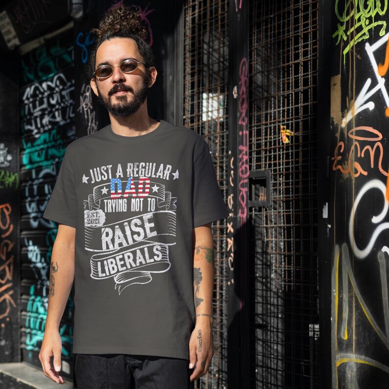 Op de afbeelding: Donkergrijs t-shirt met de witte tekst "JUST A REGULAR DAD TRYING NOT TO RAISE LIBERALS", met een Amerikaans vlagontwerp. Het shirt bevat "EST. 2021". De persoon draagt een zonnebril en staat voor een graffiti-muur.