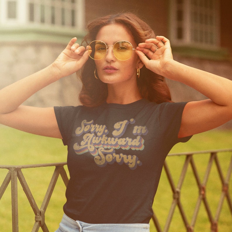 Puede incluir: Una mujer con una camiseta azul oscuro con un gr&aacute;fico de estilo retro que dice "Lo siento, soy torpe Lo siento". Lleva gafas de sol redondas amarillas y tiene el pelo largo y casta&ntilde;o.