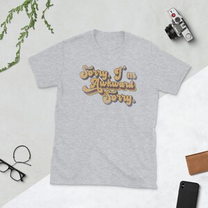 Puede incluir: Una camiseta gris jaspeada con un gr&aacute;fico de estilo retro que dice "Lo siento, soy torpe Lo siento."