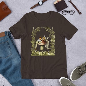 K&ouml;nnte beinhalten: Braunes T-Shirt mit einer fantastischen Illustration einer Fee und eines Pilzes. Das Kunstwerk ist von einem botanischen Rahmen mit gr&uuml;nen und gelben Akzenten umgeben. Das Design beinhaltet eine Fee mit Fl&uuml;geln und einen Pilz mit rotem Hut.