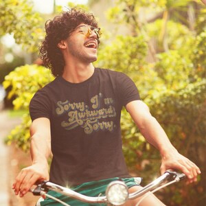 Puede incluir: Un hombre con una camiseta marr&oacute;n con el texto "Sorry, I'm Awkward Sorry" en una fuente retro. Est&aacute; montando en bicicleta y sonriendo.