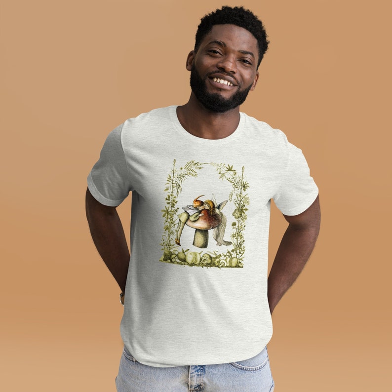 K&ouml;nnte beinhalten: Hellgraues T-Shirt mit einer Fantasy-Illustration einer Fee auf einem Pilz, umgeben von gr&uuml;nen Bl&auml;ttern. Das Design verwendet Braun-, Orange- und Gr&uuml;nt&ouml;ne. Das T-Shirt besteht aus einem weichen Material.