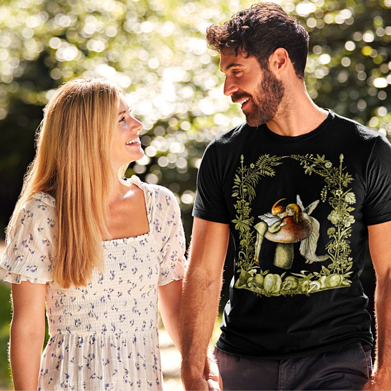 K&ouml;nnte beinhalten: Schwarzes T-Shirt mit einer skurrilen Illustration eines Feenpaares auf einem Pilz, umrahmt von einem floralen Bogen. Das Design verwendet Gr&uuml;n-, Braun- und Beiget&ouml;ne. Das Paar h&auml;lt ein Buch. Das T-Shirt wird von einer Person getragen.