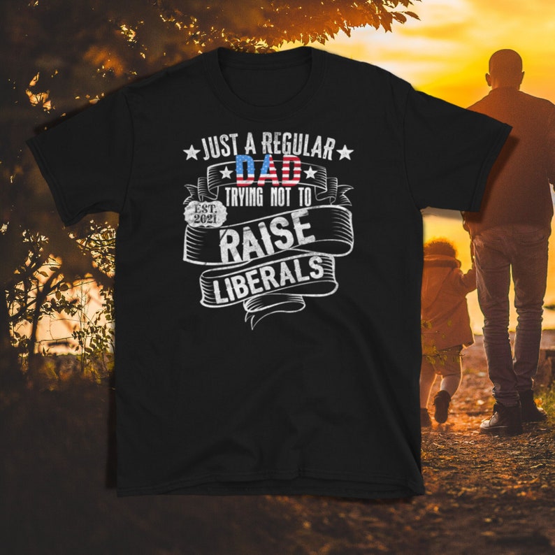 Op de afbeelding: Zwart t-shirt met de witte en rode tekst "JUST A REGULAR DAD trying not to RAISE LIBERALS". Het woord "DAD" heeft een rood-wit-blauw vlagontwerp. Het shirt heeft ook de tekst "EST. 2021".