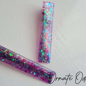 May include: Two purple hair clips with iridescent glitter embedded in the resin. The glitter is a mix of blue, green, and pink.