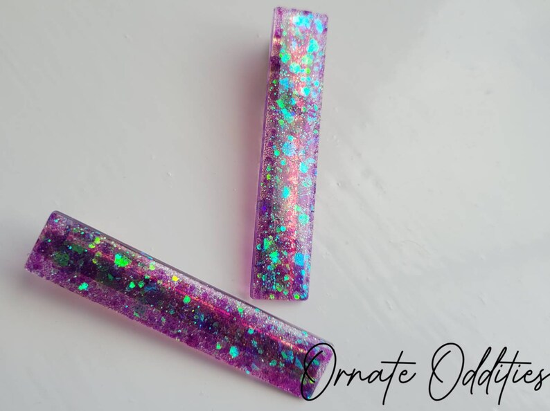 May include: Two purple hair clips with iridescent glitter embedded in the resin. The glitter is a mix of blue, green, and pink.