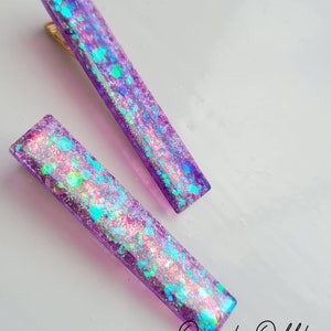May include: Two purple hair clips with iridescent glitter embedded in the resin. The clips are rectangular and have a slight curve to them.