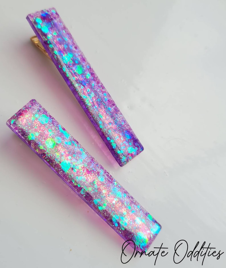May include: Two purple hair clips with iridescent glitter embedded in the resin. The clips are rectangular and have a slight curve to them.