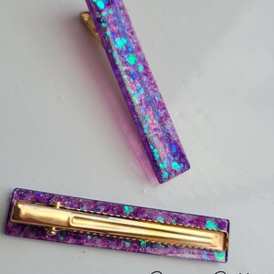 May include: Two purple hair clips with gold metal clasps. The clips are filled with iridescent glitter.