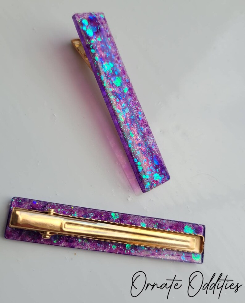 May include: Two purple hair clips with gold metal clasps. The clips are filled with iridescent glitter.