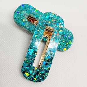 May include: Two turquoise blue hair clips with gold metal clasps. The clips are covered in iridescent glitter.