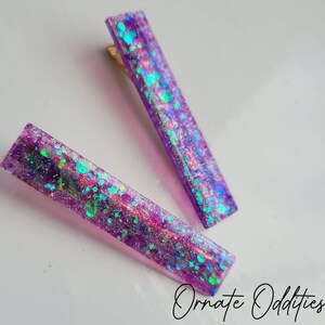 May include: Two purple hair clips with iridescent glitter embedded in the resin. The glitter is a mix of silver, green, and blue.
