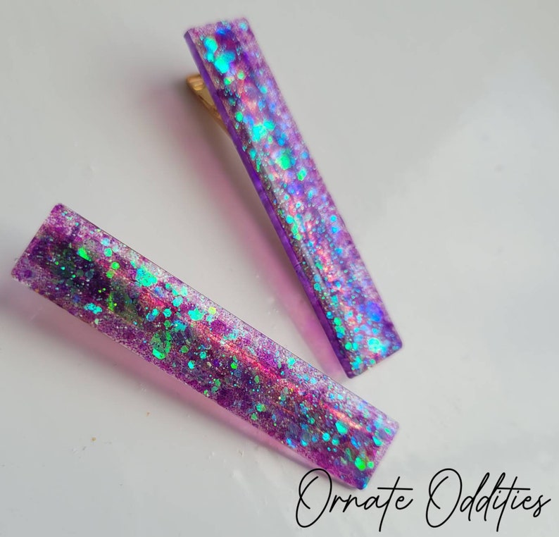May include: Two purple hair clips with iridescent glitter embedded in the resin. The glitter is a mix of silver, green, and blue.