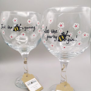 May include: Two clear glass gin balloons decorated with hand-painted white and red flowers and yellow and black bees. One glass features the text Just the ginning! and the other Let the party gin! Each glass has a brown paper tag tied to the stem.