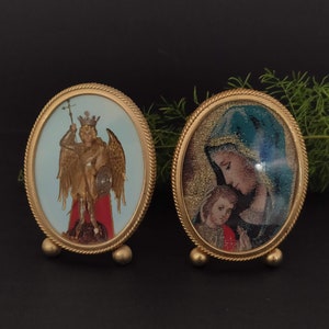 Puede incluir: Dos iconos religiosos ovalados en marcos dorados. Uno representa un ángel con corona, alas y escudo. El otro muestra a una madre y un niño. Ambos tienen un borde decorativo en forma de cuerda y pequeños pies de bola.
