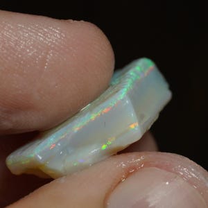 May include: A raw opal gemstone, held between fingers, shows a milky white base colour with iridescent flashes of green, red, and yellow. The stone's surface is slightly uneven, revealing its natural, unpolished state.