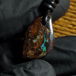 Puede incluir: Un colgante de ópalo boulder pulido con una matriz de piedra de hierro marrón oscuro que presenta destellos iridiscentes vibrantes de color azul, verde y naranja. La piedra está perforada en la parte superior y colgada de un collar de cuero negro.