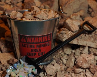 Può includere: Un piccolo secchio di metallo pieno di rocce e una pala nera piena di opali. Il secchio ha un'etichetta di avvertimento rossa con la scritta "WARNING ACTIVE MINING AREA KEEP OUT". La scena è ambientata su un letto di rocce.