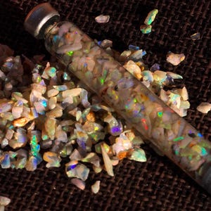 May include: A glass vial filled with iridescent opal chips, resting on a pile of similar opal fragments. The vial has a silver cap. The opals show flashes of green, blue, and orange.
