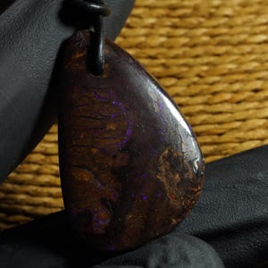 Puede incluir: Un colgante de ópalo boulder pulido con una matriz de piedra de hierro marrón oscuro que presenta destellos iridiscentes púrpuras y azules en toda la piedra. La gema en forma de lágrima cuelga de un cordón negro simple y tiene una superficie lisa.