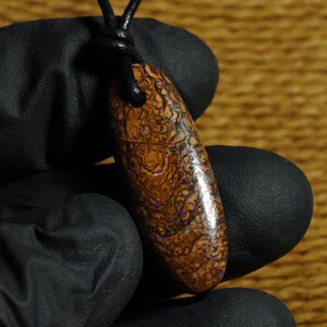 Puede incluir: Un colgante de ópalo boulder pulido y alargado con una matriz marrón compleja y arremolinada, con destellos vibrantes de color iridiscente púrpura y azul. La piedra está suspendida de un sencillo cordón de cuero negro de textura natural.