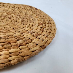 May include: Close-up of a round, woven placemat made from natural, light brown fibers. The placemat has a textured surface with a tightly woven pattern. The edges are slightly curved, and the background is white.