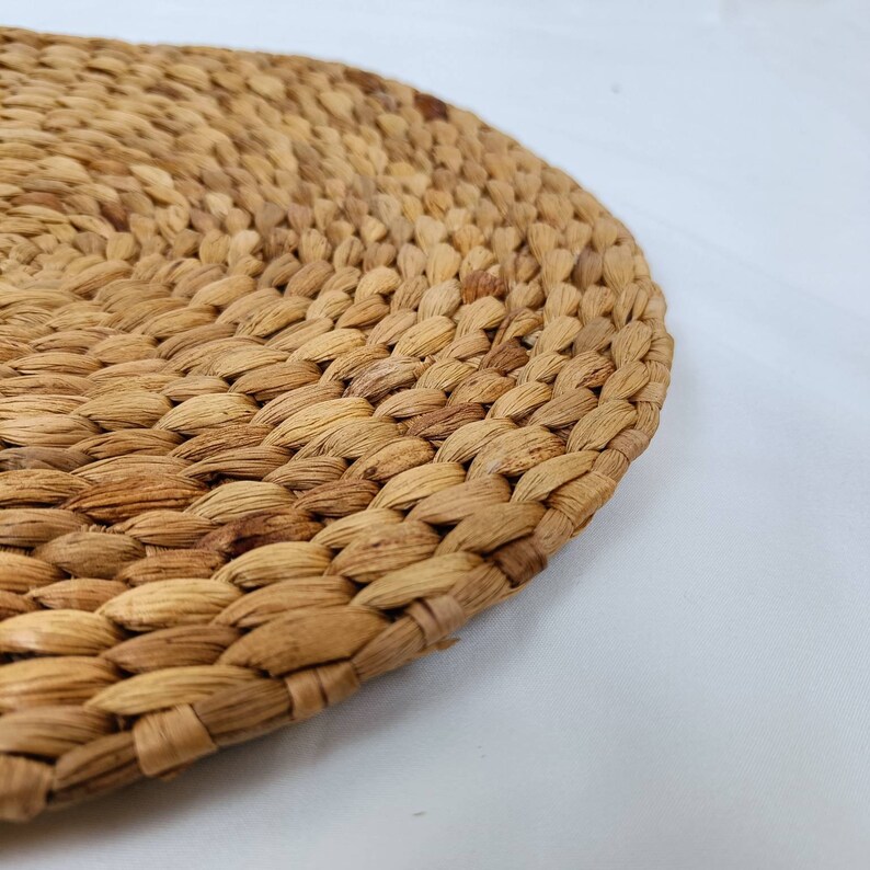 May include: Close-up of a round, woven placemat made from natural, light brown fibers. The placemat has a textured surface with a tightly woven pattern. The edges are slightly curved, and the background is white.