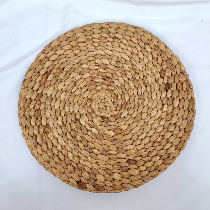 May include: A round, woven placemat made of natural-colored water hyacinth. The placemat is circular, with a spiral pattern radiating from the center. The woven texture is visible, and the overall appearance is rustic and organic.