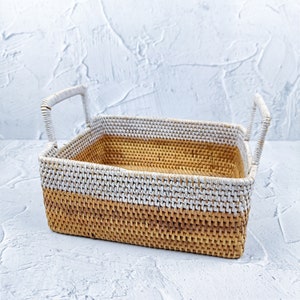 May include: A woven rectangular basket with two handles. The basket is made of natural materials and has a white and brown color scheme.