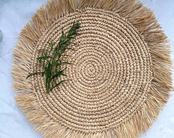 May include: A round, woven placemat made of natural fibers, featuring a fringed edge. The placemat has a spiral design in the center and is adorned with a sprig of green rosemary. The neutral color palette makes it suitable for various table settings.