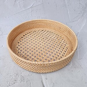 May include: A round, woven rattan tray with a lattice design in the center. The tray is light brown and has a raised edge. The tray is likely used for serving or display.
