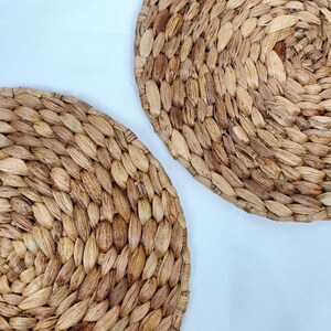 May include: Two round, woven placemats made of natural-colored, dried plant fibers. The placemats have a textured surface and are arranged diagonally against a white background. The woven design creates a rustic, organic aesthetic.
