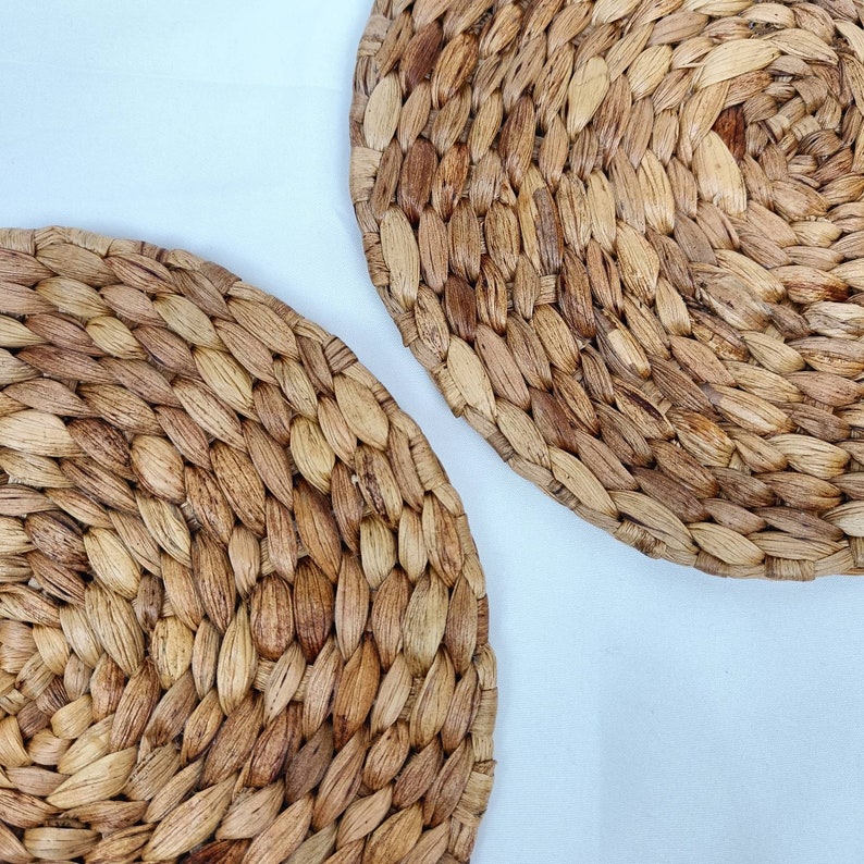 May include: Two round, woven placemats made of natural-colored, dried plant fibers. The placemats have a textured surface and are arranged diagonally against a white background. The woven design creates a rustic, organic aesthetic.
