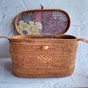 May include: Open, rectangular woven rattan handbag with a patterned fabric lining. The bag is a natural brown color, with a decorative woven pattern on the front. The interior lining features a floral and geometric design in shades of purple, green, and blue.