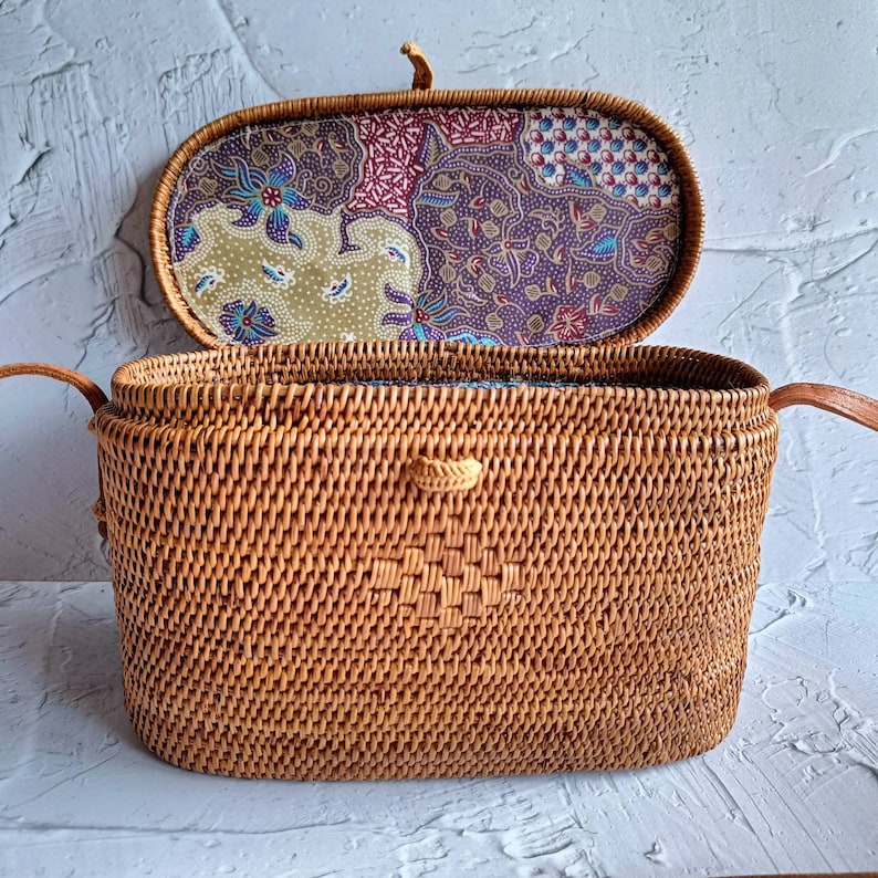 May include: Open, rectangular woven rattan handbag with a patterned fabric lining. The bag is a natural brown color, with a decorative woven pattern on the front. The interior lining features a floral and geometric design in shades of purple, green, and blue.