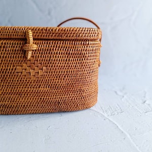 May include: A woven rattan handbag in a warm brown hue. The rectangular bag features a woven pattern, a handle, and a decorative closure. The bag is set against a light gray background, highlighting its texture and design. A stylish accessory.