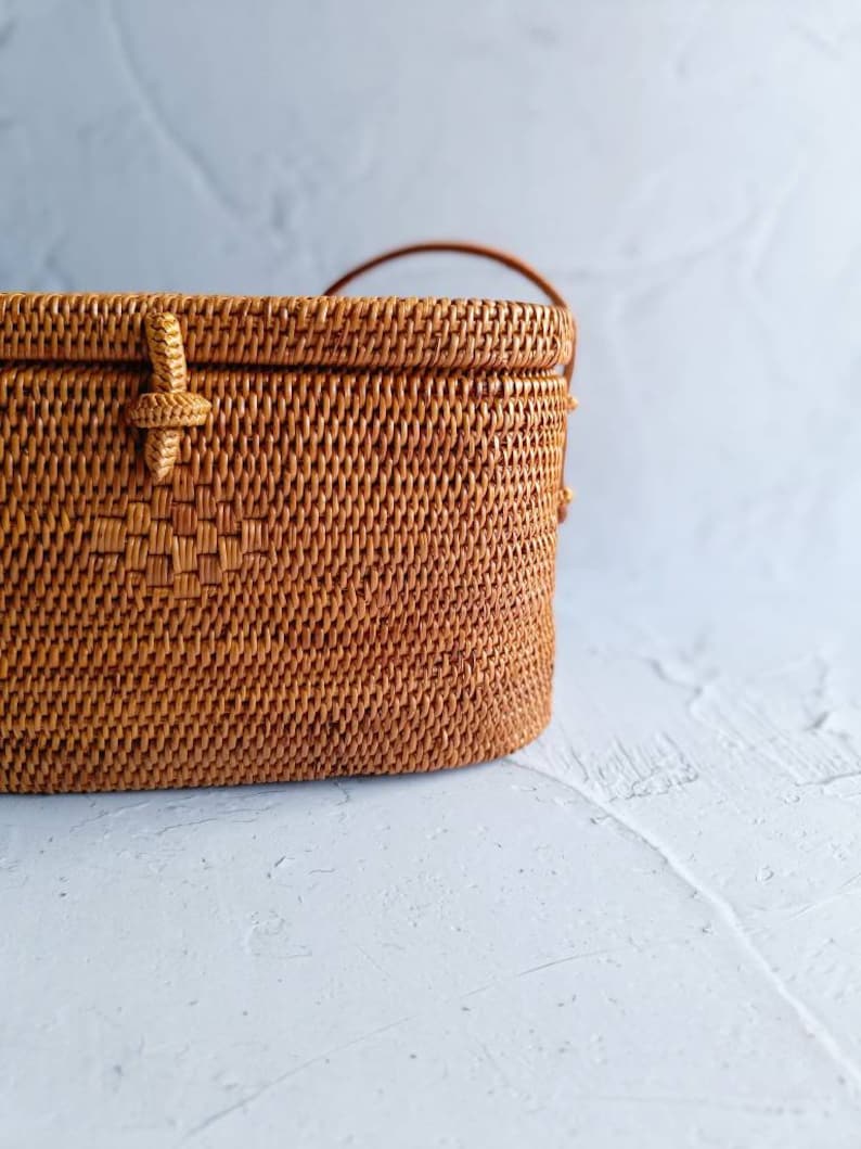 May include: A woven rattan handbag in a warm brown hue. The rectangular bag features a woven pattern, a handle, and a decorative closure. The bag is set against a light gray background, highlighting its texture and design. A stylish accessory.