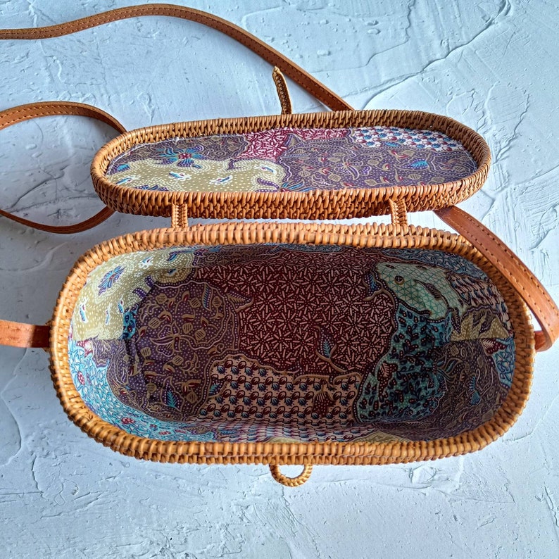 May include: Open oval-shaped rattan handbag with a patterned fabric lining. The bag has a long, adjustable strap and a woven exterior. The interior fabric features a mix of colors including red, green, and blue.
