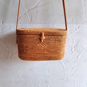 May include: A rectangular, woven rattan crossbody bag with a light brown leather strap. The bag features a woven pattern and a decorative clasp. The bag is designed to be worn over the shoulder.