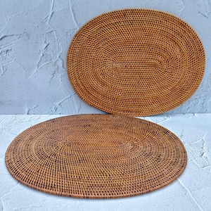 Può includere: Due tovagliette ovali in rattan intrecciato. Le tovagliette sono realizzate in rattan marrone naturale e hanno un motivo intrecciato.