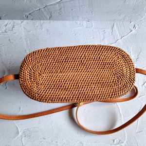 May include: A woven rattan oval-shaped handbag with a brown leather strap. The bag is a light brown color and has a textured surface. The strap is a lighter shade of brown and is attached to the sides of the bag.