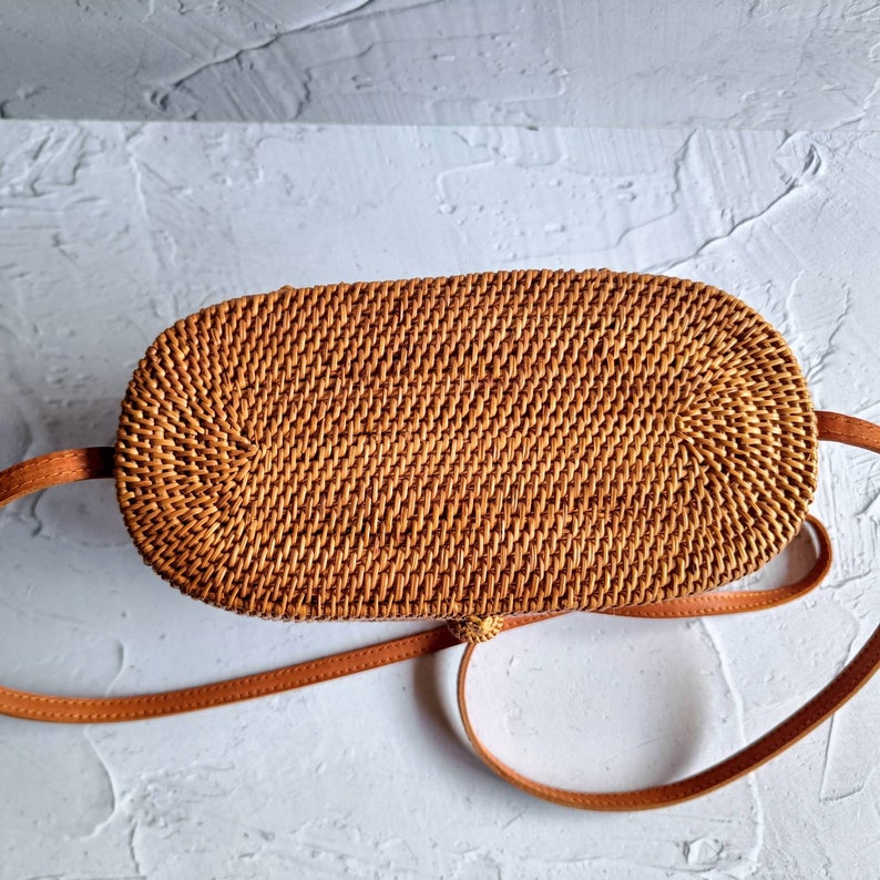 May include: A woven rattan oval-shaped handbag with a brown leather strap. The bag is a light brown color and has a textured surface. The strap is a lighter shade of brown and is attached to the sides of the bag.