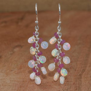Peut inclure: Boucles d'oreilles pendantes composées d'opales blanches et de petites pierres roses rondes. Les pierres sont enfilées sur des chaînes et des crochets argentés. Les boucles d'oreilles sont présentées sur un fond en bois.